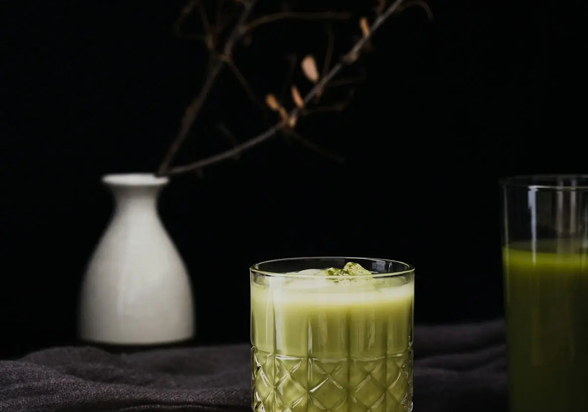 a glass of green juice next to a wooden cutting board