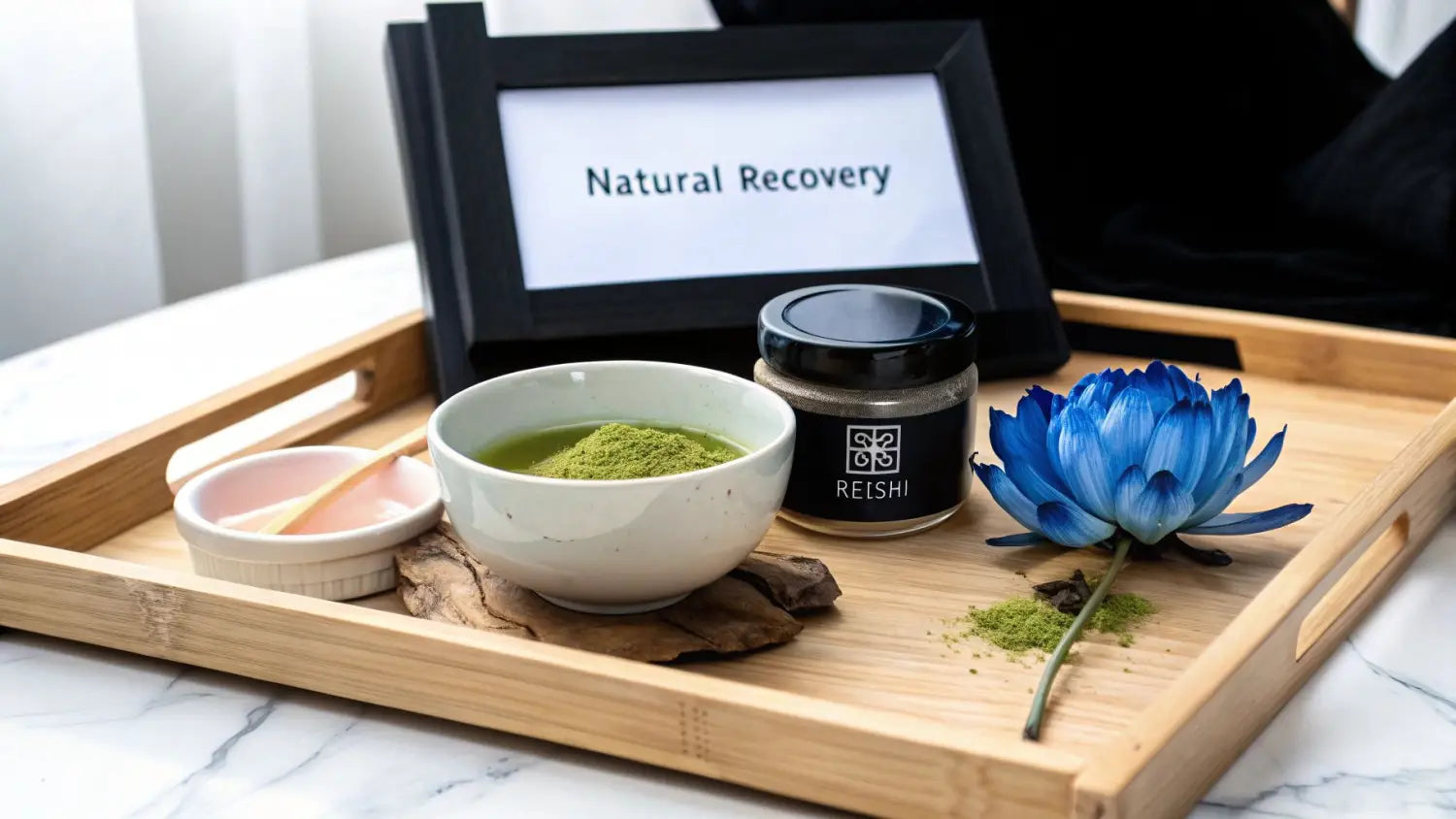 A wooden tray with a bowl of green powder, a 'REISHI' jar, a blue flower, and a 'Natural Recovery' sign.