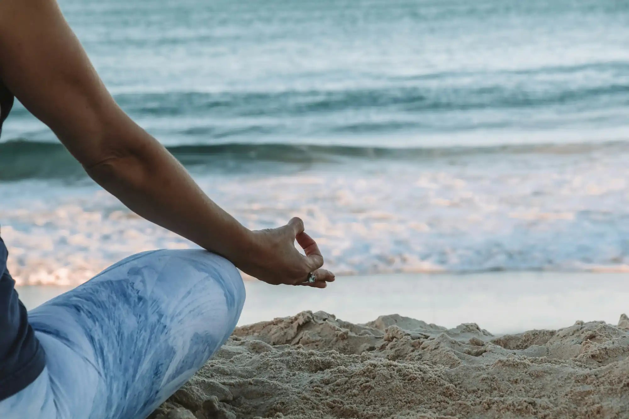 Hand in a meditation pose resting on a leg while sitting on sand.