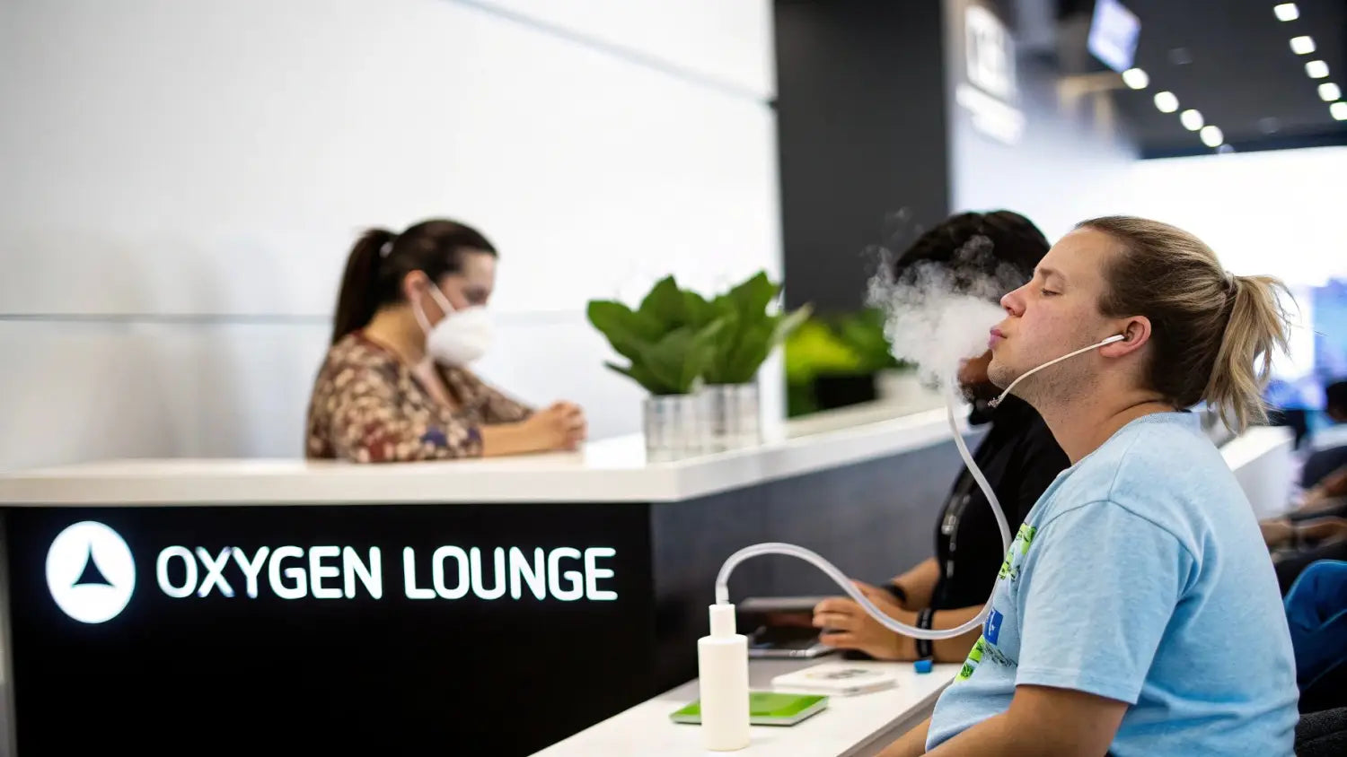 A man exhales vapor from an oxygen therapy device in an Oxygen Lounge, with a masked woman in the background.