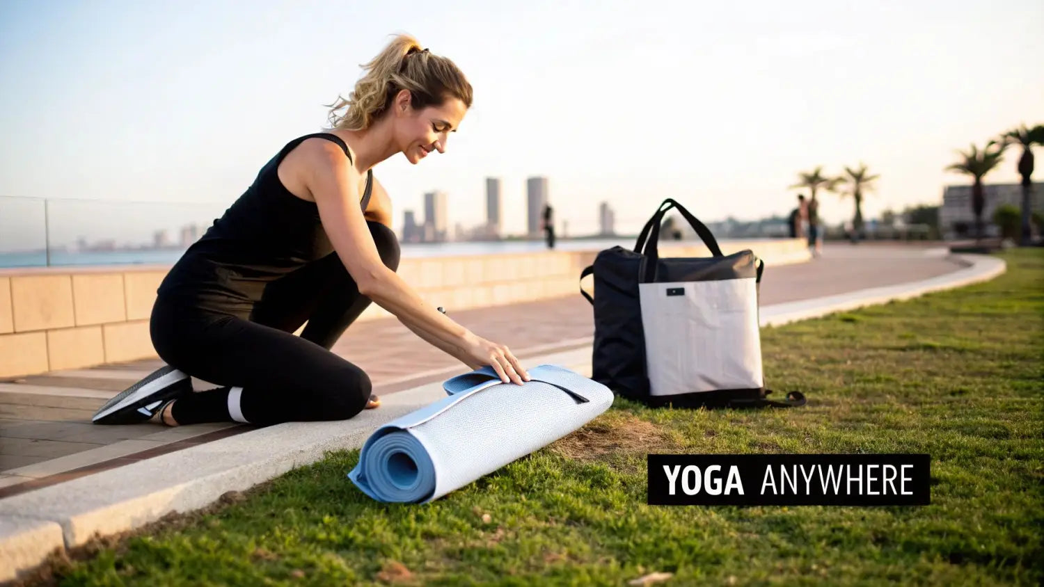 A smiling woman prepares her light blue foldable yoga mat on grass outdoors by the water.