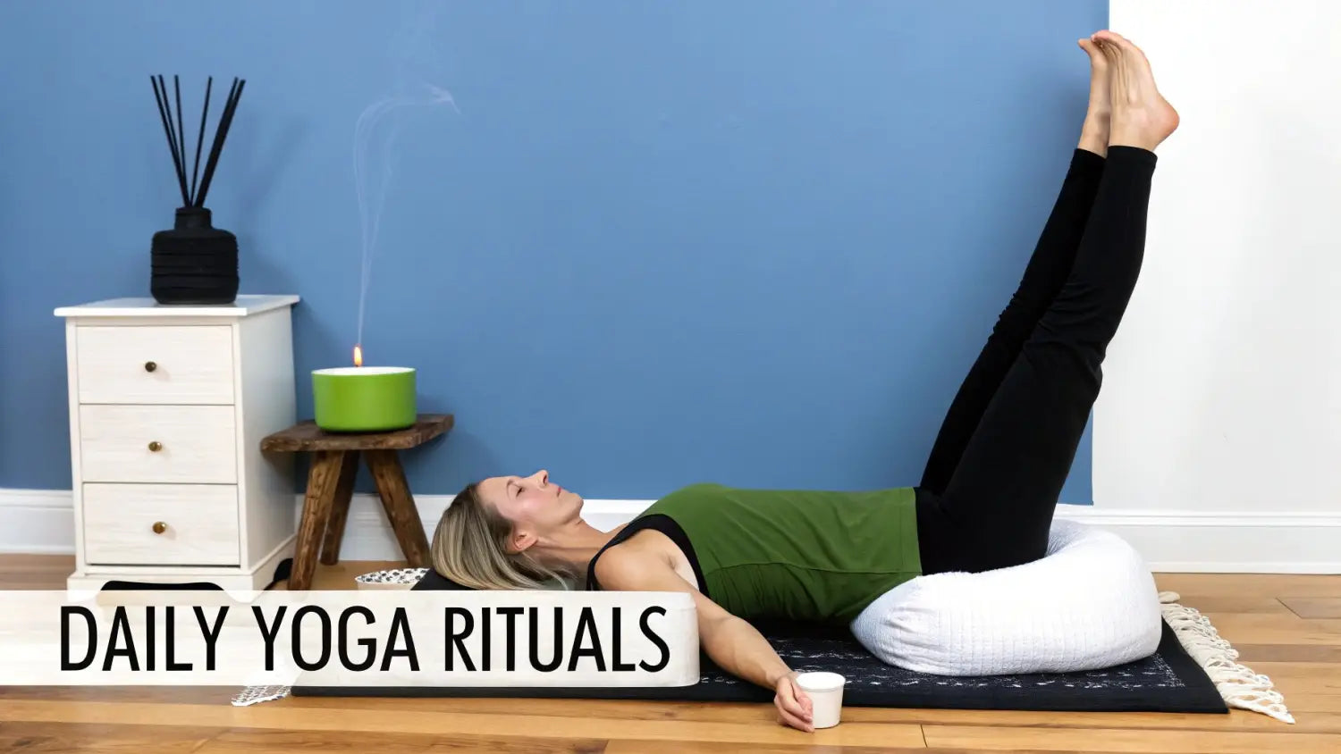 A woman performs a yoga pose (legs-up-the-wall) on a mat with a bolster, next to a lit candle and diffuser.